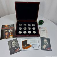 Overview of mahogany presentation box open showing twelve coins, brochure, green apple for scale, and certificate
