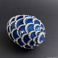 Blue enamel decorative egg with scalloped pattern and brilliant stones on black background, shown from an angle.