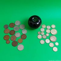 Wide shot of all coins including Canadian and British coins arranged around a black spherical object for scale, on a green background.