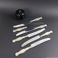Full set of six knives with stainless steel blades and ornate sterling silver handles on black background, shown with black decorative round object for scale.