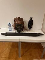 Full view of the lot with both masks and long carved sculpture displayed on a white table with a water bottle for scale.