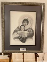 Full frontal image of the framed charcoal print artwork depicting an adult in fur-lined hood holding a baby, showing the entire piece and frame.