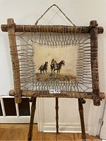 View of entire framed animal hide painting hanging, showing cowboy and two horses in rustic wood frame