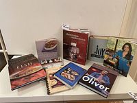 Shelf or table display of 11 cookbooks including Williams-Sonoma, Jamie Oliver, Eva Longoria, chocolates confections, Joy of Cooking, and others.