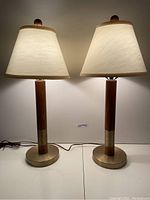 Full view of the pair of lit mid century wood and brass lamps with conical fabric shades, showing height and overall design.