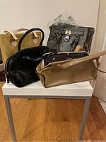 Four handbags on a table including green patent tote, black dome satchel, gray studded tote, and camel shoulder bag