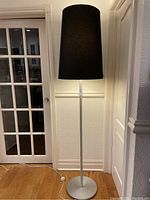 Full view of the tall floor lamp with black shade illuminated against wall and door in background