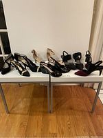 Five pairs of brand new ladies shoes displayed on two side-by-side white tables on wooden floor, showing variety in styles and colors including black and burgundy.