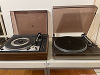 Two vintage stereo turntables side by side on a white surface with their plastic dust covers open, showing the Dual 1225 on the left with wood-tone base and the Dual DCS 5025P on the right with silver metal case.