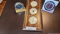 Photo of a wall-mounted weather station with three round dials labeled barometer, thermometer, and hygrometer, mounted on a wooden panel with gold trim, alongside a small round blue-faced clock with a gold-tone base, both resting on a table.