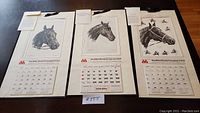 Three calendars showing detailed black and white horse portraits with calendar grids, mounted on cream paper with black hanging ribbons and written information.
