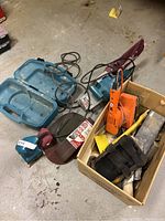 Entire lot showing Makita belt sander with dust bag, Black & Decker orange belt sander, a Makita palm sander, sanding belts, sanding sheets, and carrying case on concrete floor.