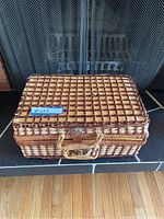 Closed vintage rectangular woven wicker picnic basket with a brown and tan checkered pattern, with handle and clasps visible, placed on a dark surface with a fireplace screen in the background.