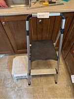 Photo showing the foldable two-step stool next to the small white plastic stool on a tiled floor near kitchen cabinetry.