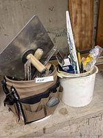 Fabric tool bag filled with various tiling hand tools including hawk, spreader, and trowels.