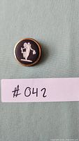 Front view of Wedgwood dress pin showing white classical figure on black background with gold-tone edge.