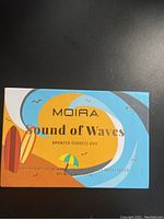 Closed box with colorful wave and beach design, 'Moira Sound of Waves Bronzed Goddess Duo' branding.