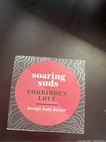 Top view of boxed Soaring Suds Forbidden Love Mango Body Butter with red circular label on black and gray wavy pattern box.
