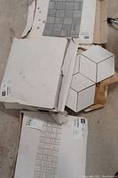 Photo of several boxed mosaic tile sheets showing tiles with light gray small square pattern, white hexagon shapes, and a white 3D textured rectangular pattern.