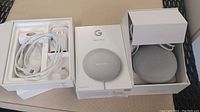 Two Google Nest Mini speakers shown, one boxed, one out of box with packaging and accessories inside