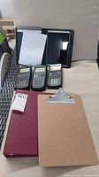 Photo showing three Texas Instruments calculators (two TI-89 and one TI-83 Plus), an HP tablet, two clipboards, and a maroon binder.