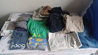 Various men's pants and t-shirts folded and laid out on box. Pants include blue denim, khaki, black, and tan colored pants. T-shirts in different shades including green, gray with prints.