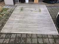 Full view of the outdoor area rug laid out on stone pavement showing wear, dirt and a central damaged area.