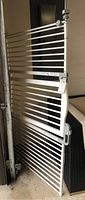 Full view of a white safety gate showing horizontal bars, mounting hardware, and a handle release mechanism.