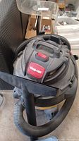 Side and top view of the Shop-Vac showing the logo, power switch, and attached hose with wide nozzle.