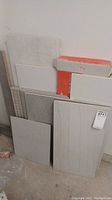Assortment of various floor and wall tiles including large rectangular tiles with wood grain texture, smooth white tiles, and other gray toned tiles with different surface finishes.
