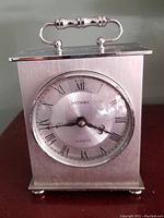 Front view of Metamec stainless steel carriage clock showing dial with Roman numerals and clock hands, brushed steel body and handle