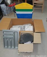 Four triangular plastic bins stacked in colors yellow, green, white, and blue; gray cutlery tray and black lid of rubbish pail in cardboard boxes.
