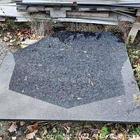 Photo showing a black granite countertop corner piece with polished surface and natural granite patterns, placed outdoors on concrete with some debris around.