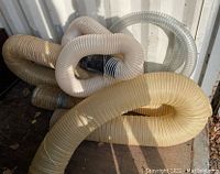 Photo of multiple assorted flexible hoses coiled and looped on ground. Colors vary from beige, off-white to translucent. Corrugated plastic construction visible.