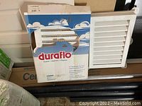 Photo showing two Duraflo gable vents, one outside and one presumably in the box, white with horizontal slats, packaging box with blue and white sky/cloud design and product specs.