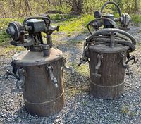 Photo showing three used metal paint pressure pots outdoors on gravel surface with hoses and attached pressure regulators. Pots have rust and wear marks, lids secured with clamps.