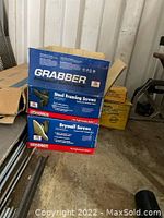 Photo showing two cardboard boxes on the floor; one is labeled 'Grabber Professional Quality Screws Steel Framing Screws' in blue, and the other is labeled 'Grabber Drywall Screws' in red.