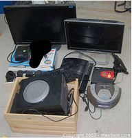 Photo showing two LED monitors, a PlayStation 3 console, Logitech X530 speakers with subwoofer, a red and black Motomaster Eliminator power supply, a silver CD/radio clock radio, and assorted cables and peripherals on the floor.