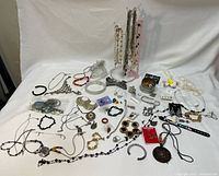 Overview of assorted costume jewelry including necklaces, bracelets, rings, and earrings spread on a white surface with an empty jewelry stand to the side.