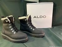 Pair of black ALDO combat winter boots shown with white ALDO box in background on dark fabric surface.