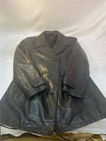 Black faux leather trench coat laid flat showing front with double-breasted buttons, wide collar, long sleeves, and zippered pockets.