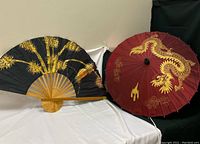 Photo of both the traditional Thai folding fan and large umbrella displayed together on a fabric-covered surface.