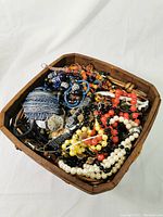 Wicker basket filled with assorted costume jewelry, including various colored beaded necklaces and watches, on a white background.