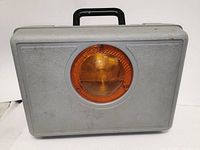 Closed gray plastic carrying case with an amber circular light centered on the front, showing surface wear and minor scuffs.