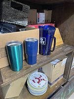 Blue Bud Light plastic pitcher alongside green and blue insulated tumblers on wooden shelf.