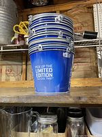 Stack of fifteen blue metal beer buckets with silver rims and handles. One bucket shows white promotional text 'PICK UP THE LIMITED EDITION 16oz COOL TWIST'.