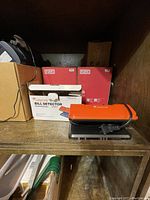 Photo of the brand new electronic bill detector device with orange top cover, placed on a shelf alongside other boxed items.
