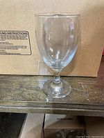 Single clear stemmed beverage glass placed on wooden surface in front of a cardboard box.