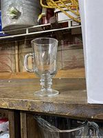 Single clear glass espresso mug with ridged ring stem on a wooden surface, background blurred with other items.