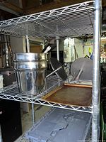 Photo showing two stainless steel professional condiment storers and servers placed on a metal wire shelf, each with a hinged handle-lid and polished finish in a commercial kitchen setting.